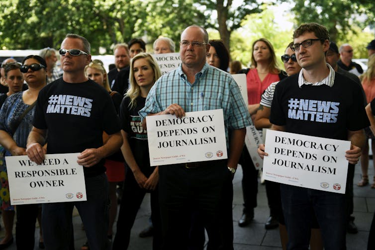 Three men hold signs saying 'democracy depends on journalism' as they protest against policies at The Denver Post newspaper.